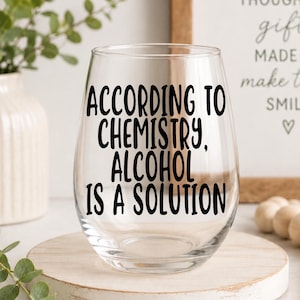 May include: A clear stemless wine glass featuring the black printed text ACCORDING TO CHEMISTRY, ALCOHOL IS A SOLUTION. The glass has a rounded bowl shape with a smooth rim and a thick base, designed for holding beverages in a standard drinking capacity.