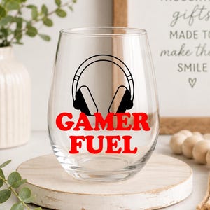May include: A clear stemless wine glass with the words "GAMER FUEL" in red and a black headphone graphic. The glass sits on a round wooden stand. A sign in the background reads "THOUGHTFUL gifts MADE TO make them SMILE."