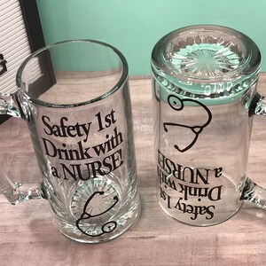 May include: Two clear glass beer mugs with handles. One mug is upright, the other is upside down. Both mugs have the text "Safety 1st Drink with a NURSE!" and a stethoscope graphic in black. The mugs are on a light wood-grain surface.