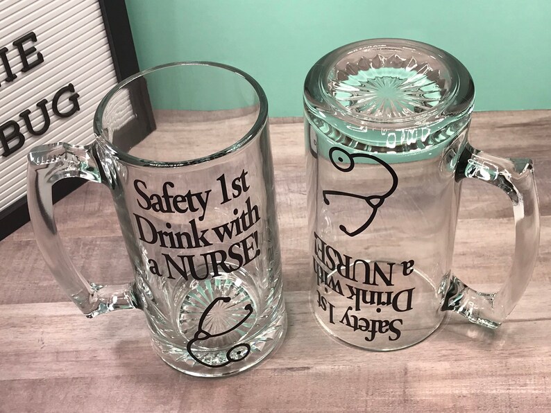 May include: Two clear glass beer mugs with handles. One mug is upright, the other is upside down. Both mugs have the text "Safety 1st Drink with a NURSE!" and a stethoscope graphic in black. The mugs are on a light wood-grain surface.