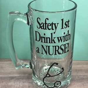 May include: A clear glass beer mug with a handle. The mug features the text "Safety 1st Drink with a NURSE!" in black, along with a stethoscope graphic. The mug has a decorative base.