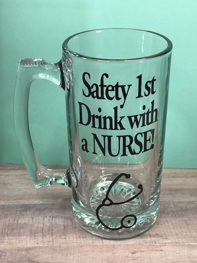 May include: A clear glass beer mug with a handle. The mug features the text "Safety 1st Drink with a NURSE!" in black, along with a stethoscope graphic. The mug has a decorative base.