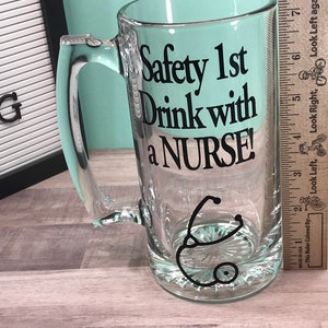 May include: Clear glass beer mug with a handle. The mug features the text "Safety 1st Drink with a NURSE!" in black, along with a black stethoscope graphic. The mug is on a wooden surface.