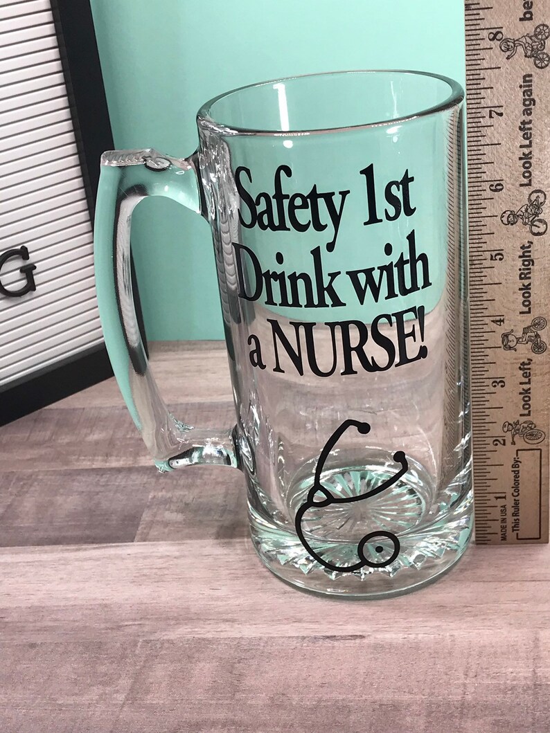 May include: Clear glass beer mug with a handle. The mug features the text "Safety 1st Drink with a NURSE!" in black, along with a black stethoscope graphic. The mug is on a wooden surface.
