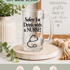 May include: A clear glass beer mug with a handle, featuring the text "Safety 1st Drink with a NURSE!" in black, along with a stethoscope graphic. The mug is displayed on a wooden coaster, with decorative elements in the background.