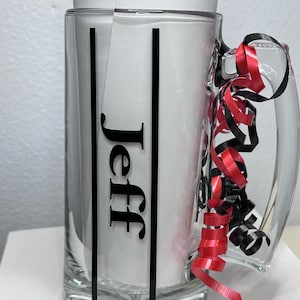 May include: A clear glass beer mug with a handle, featuring the name "Jeff" in black lettering. The mug is decorated with black vertical stripes and red and black curling ribbon. The mug is empty.