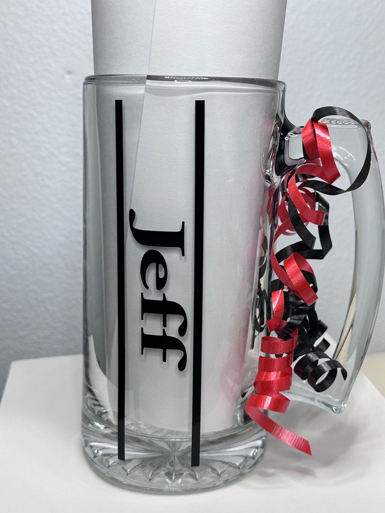 May include: A clear glass beer mug with a handle, featuring the name "Jeff" in black lettering. The mug is decorated with black vertical stripes and red and black curling ribbon. The mug is empty.