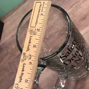 May include: Clear glass beer mug with a handle. The mug has the words "Safety Drink a NU" printed on the side. A wooden ruler is placed inside the mug, measuring approximately 6 inches in height.