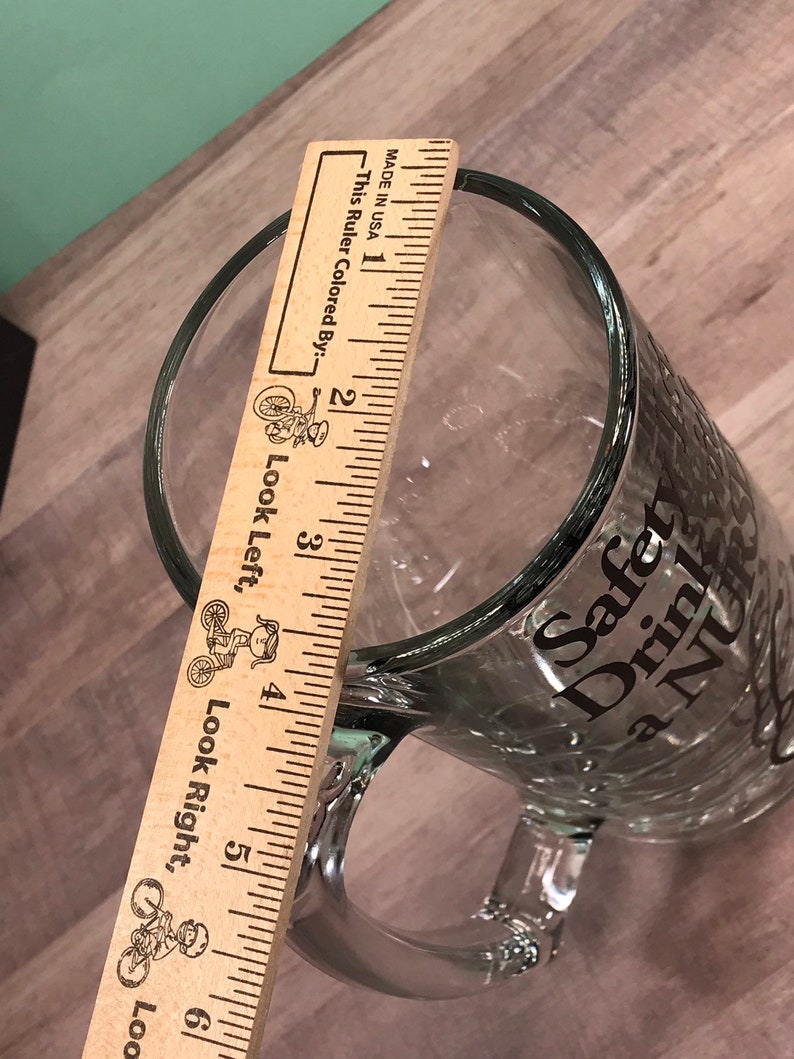 May include: Clear glass beer mug with a handle. The mug has the words "Safety Drink a NU" printed on the side. A wooden ruler is placed inside the mug, measuring approximately 6 inches in height.
