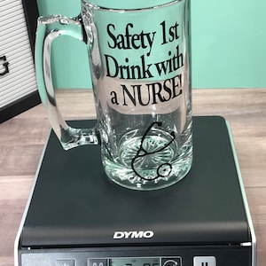 May include: A clear glass beer mug with a handle, featuring the text "Safety 1st Drink with a NURSE!" in black. A stethoscope graphic is below the text. The mug is on a black digital scale, weighing 2.85 lbs.
