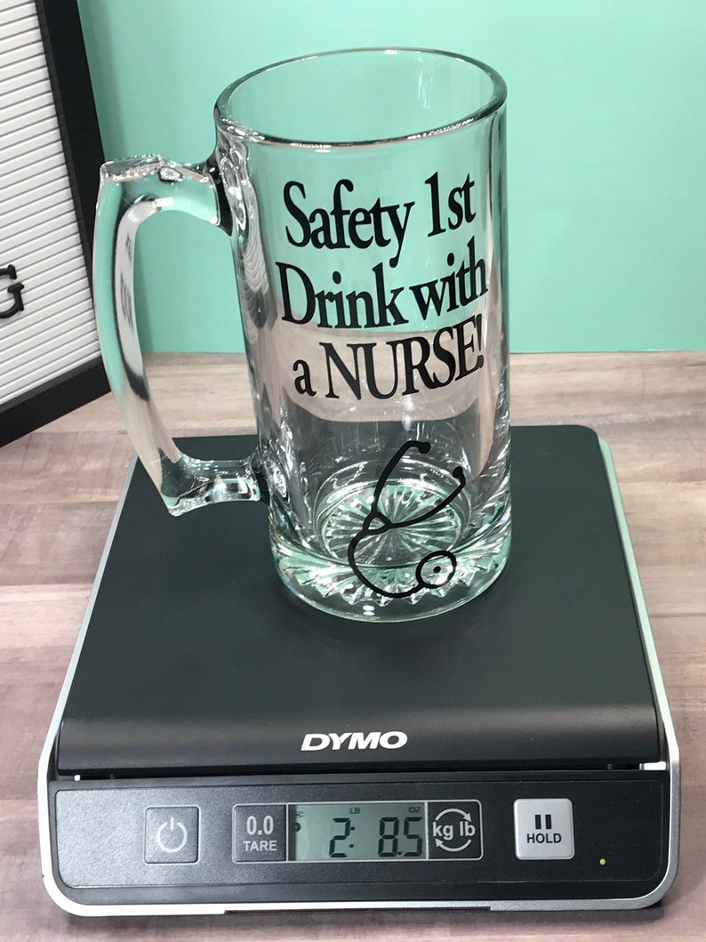 May include: A clear glass beer mug with a handle, featuring the text "Safety 1st Drink with a NURSE!" in black. A stethoscope graphic is below the text. The mug is on a black digital scale, weighing 2.85 lbs.