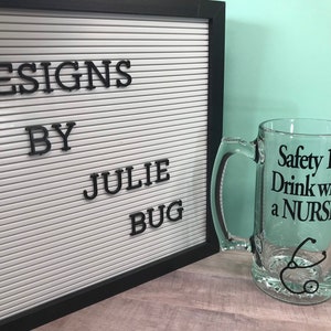 May include: A clear glass beer mug with a handle, featuring the text "Safety 1st Drink with a NURSE!" in black. A black stethoscope graphic is below the text. A white letter board with the text "DESIGNS BY JULIE BUG" is in the background.
