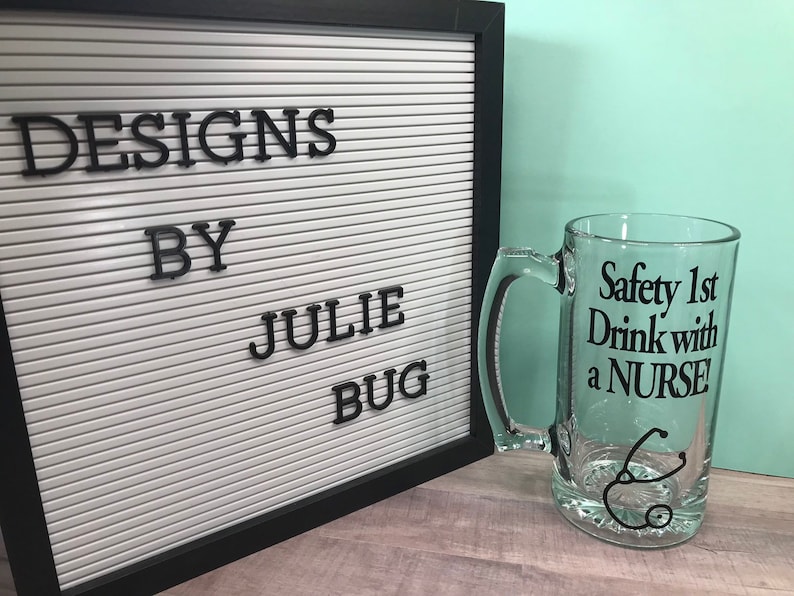 May include: A clear glass beer mug with a handle, featuring the text "Safety 1st Drink with a NURSE!" in black. A black stethoscope graphic is below the text. A white letter board with the text "DESIGNS BY JULIE BUG" is in the background.