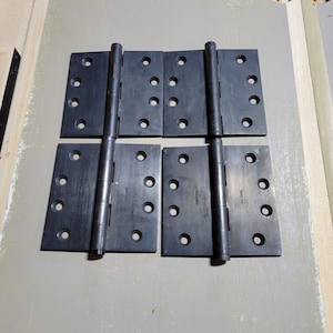 May include: Four black metal door hinges with a brushed finish. Each hinge has five round holes and a central pin.