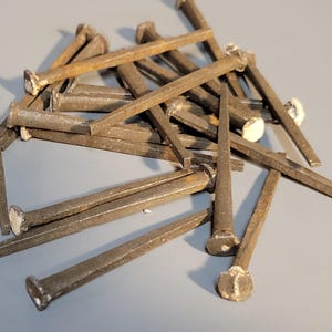 May include: A pile of antique, rusty, square-headed nails. The nails are a dark brown color with a slightly weathered appearance. The heads are a lighter color, possibly from oxidation. These nails are likely for woodworking or construction.