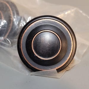 Könnte beinhalten: Ein schwarzer und kupferfarbener Schrankknopf mit konzentrischem Kreisdesign.