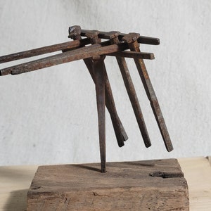 May include: A rustic metal sculpture featuring a series of angled, rusty metal rods attached to a wooden base. The sculpture has a minimalist design and a weathered appearance, suggesting an industrial or vintage aesthetic.