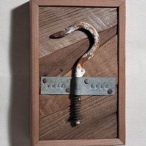Puede incluir: Una obra de arte enmarcada en madera rústica con un gancho de metal oxidado con un tornillo, una placa de metal con las palabras "WHAT EVER" estampadas en ella y un fondo de madera.
