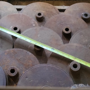 May include: A group of aged, circular metal discs with central holes, possibly industrial parts. A yellow measuring tape, marked in inches, is laid across the discs, indicating size. The discs are a rusty brown, suggesting age and exposure to the elements.