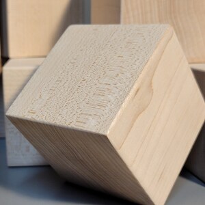 May include: Close-up of a light-coloured wooden cube with visible grain and rounded edges. Other similar cubes are visible in the background. The cube is angled, showcasing its texture and form. Suitable for crafts or decorative purposes.