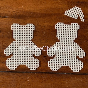 May include: Two white plastic bear shapes with a grid pattern, ready for cross-stitch. The bears are separated and there is a small white plastic hat shape in the upper right corner of the image.