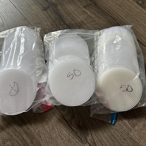May include: Three clear plastic bags each containing a stack of white, circular plastic canvas pieces. Each bag has "50" written on the canvas in black marker. The canvas pieces have a textured, perforated surface.