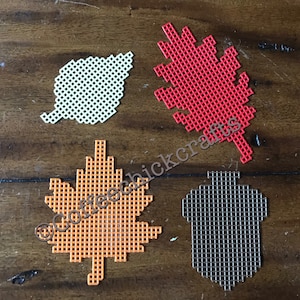 May include: Four plastic canvas shapes: a white leaf, a red leaf, an orange maple leaf, and a brown acorn. The shapes are arranged on a wooden surface. The text "coffeechickcrafts" is visible.