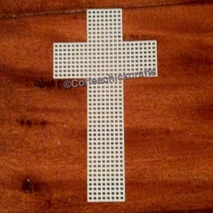 May include: A white plastic canvas cross with a grid pattern. The cross is centered on a brown wooden surface. The text "©Coffeechickcrafts" is visible on the cross.