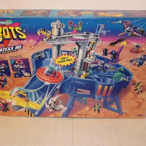May include: A Micro Machines ZBOTS Vortexx HQ playset box. The box features a blue and orange color scheme with illustrations of robots, a base, and the text "Vortexx HQ". The playset includes a base with multiple levels and a launch ramp.