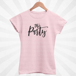 以下が含まれることがあります： 黒い筆記体で「the Party」と書かれた、薄いピンク色のTシャツ。木製のハンガーにかかっています。テキストはスタイリッシュな手書きフォントです。