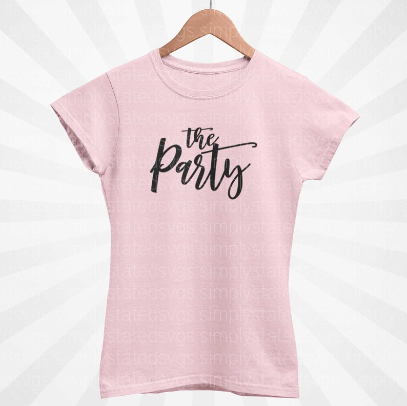 以下が含まれることがあります： 黒い筆記体で「the Party」と書かれた、薄いピンク色のTシャツ。木製のハンガーにかかっています。テキストはスタイリッシュな手書きフォントです。
