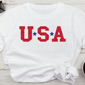 K&ouml;nnte beinhalten: Wei&szlig;es T-Shirt mit dem Wort "USA" in roten Blockbuchstaben und zwei blauen Sternen auf beiden Seiten des "S".