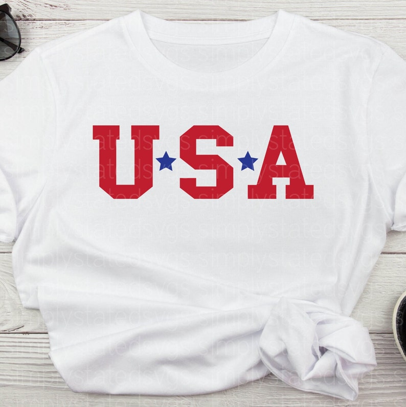K&ouml;nnte beinhalten: Wei&szlig;es T-Shirt mit dem Wort "USA" in roten Blockbuchstaben und zwei blauen Sternen auf beiden Seiten des "S".