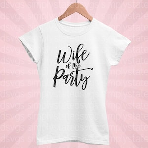 以下が含まれることがあります： 黒い筆記体で「Wife of the Party」と書かれた白いTシャツ。木製のハンガーにかけられており、背景はピンク色です。