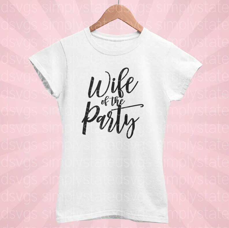 以下が含まれることがあります： 黒い筆記体で「Wife of the Party」と書かれた白いTシャツ。木製のハンガーにかけられており、背景はピンク色です。