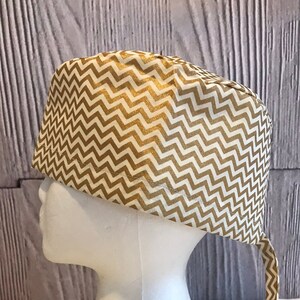 Puede incluir: Un gorro quirúrgico de frotación con estampado de chevron dorado y blanco con una corbata larga. El gorro está en una cabeza de maniquí blanca.