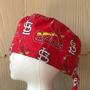 May include: Red scrub hat with a St. Louis Cardinals print featuring the team logo, a cardinal bird, and the word "Cardinals". The hat has two long ties.