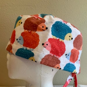 May include: A white scrub hat with a colorful hedgehog print. The hat has a red, white, and blue tie.