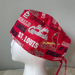 May include: Red and black surgical scrub hat with a St. Louis Cardinals theme. The hat has a tie on the back and features the team's logo and text.