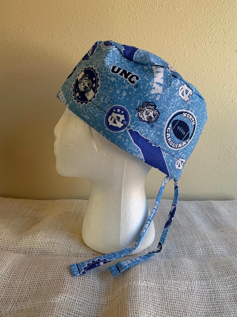 May include: A light blue and white patterned scrub cap with "UNC" lettering and various logos. The cap has a tie closure. Logos include a football, a ram, and the outline of North Carolina. The cap is displayed on a white mannequin head.