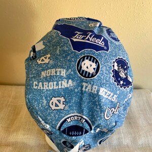 May include: A light blue and white patterned scrub cap featuring the words "North Carolina" and "Tar Heels". The cap has a tie closure and is made of a soft fabric. The cap has multiple logos and designs.