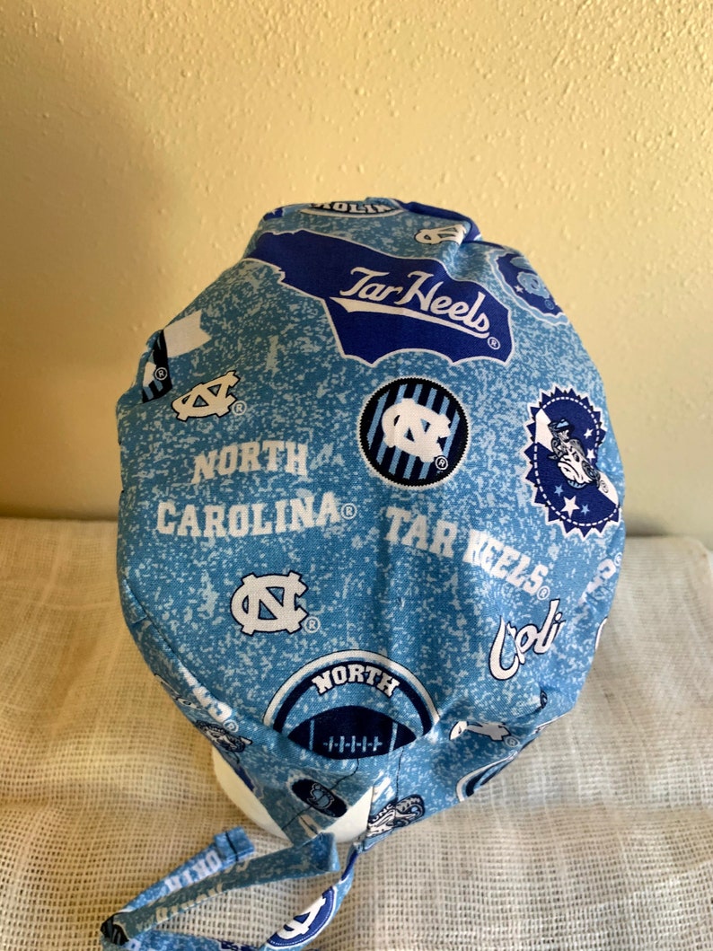 May include: A light blue and white patterned scrub cap featuring the words "North Carolina" and "Tar Heels". The cap has a tie closure and is made of a soft fabric. The cap has multiple logos and designs.
