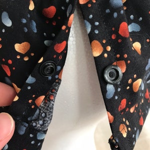 May include: Black fabric hat with a colourful paw print pattern in orange, blue, and tan. The hat has two black snap closures and a mesh lining. The hat is designed to be worn on the head.