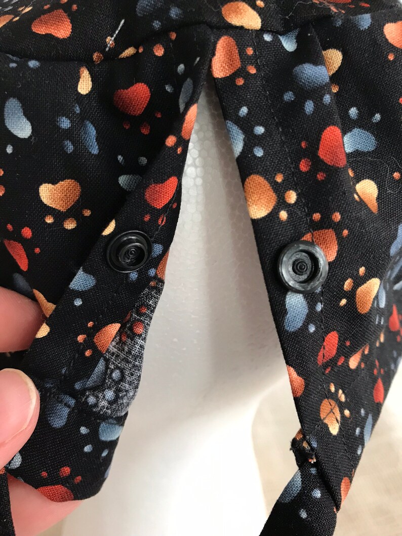 May include: Black fabric hat with a colourful paw print pattern in orange, blue, and tan. The hat has two black snap closures and a mesh lining. The hat is designed to be worn on the head.