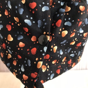 May include: Black fabric hat patterned with paw prints in orange, tan, and blue. The hat features a black button and ties. Suitable for medical or veterinary professionals, or anyone who loves animals.
