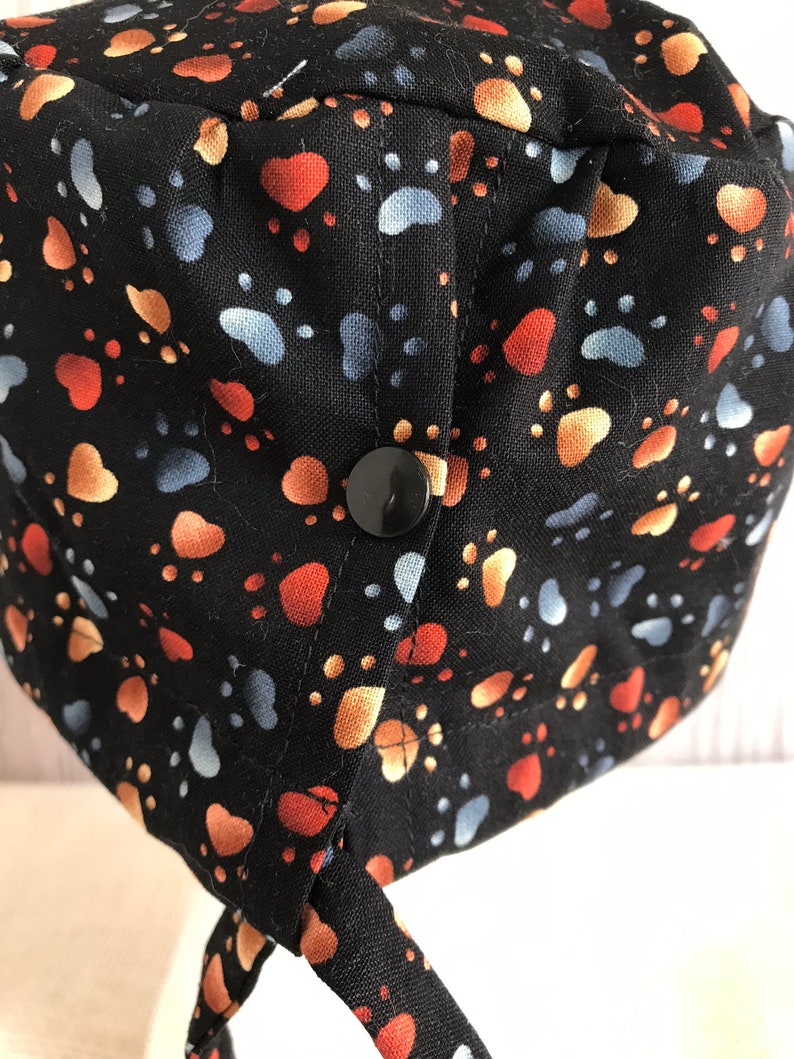 May include: Black fabric hat patterned with paw prints in orange, tan, and blue. The hat features a black button and ties. Suitable for medical or veterinary professionals, or anyone who loves animals.