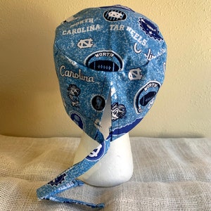 May include: A light blue and white patterned scrub cap featuring the words "North Carolina" and "Tar Heels", along with football graphics and the UNC logo. The cap is tied at the back and is displayed on a white mannequin head.