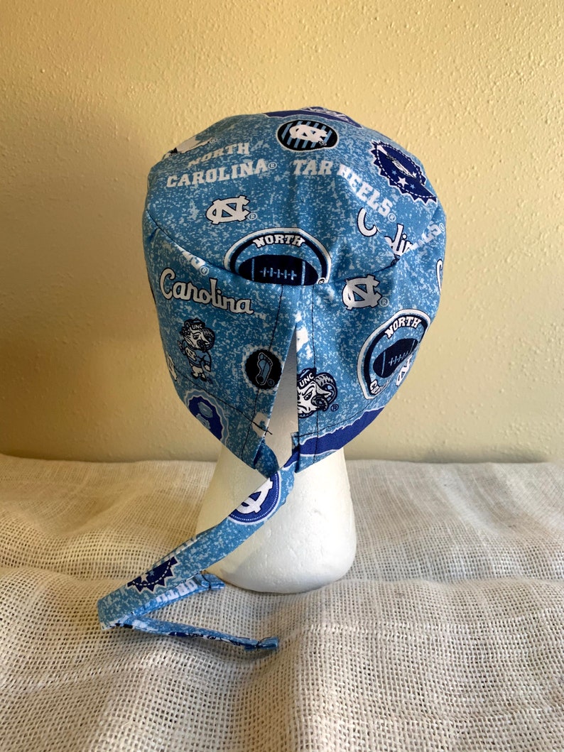 May include: A light blue and white patterned scrub cap featuring the words "North Carolina" and "Tar Heels", along with football graphics and the UNC logo. The cap is tied at the back and is displayed on a white mannequin head.