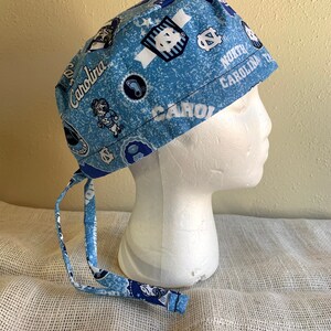 May include: A light blue scrub cap featuring a repeating pattern of Carolina logos and text. The cap has matching fabric ties at the back. The words "North Carolina" and "Carolina" are printed on the cap.