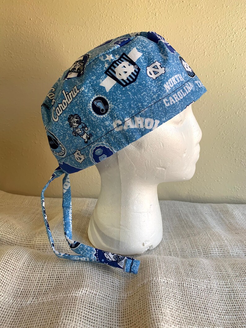May include: A light blue scrub cap featuring a repeating pattern of Carolina logos and text. The cap has matching fabric ties at the back. The words "North Carolina" and "Carolina" are printed on the cap.